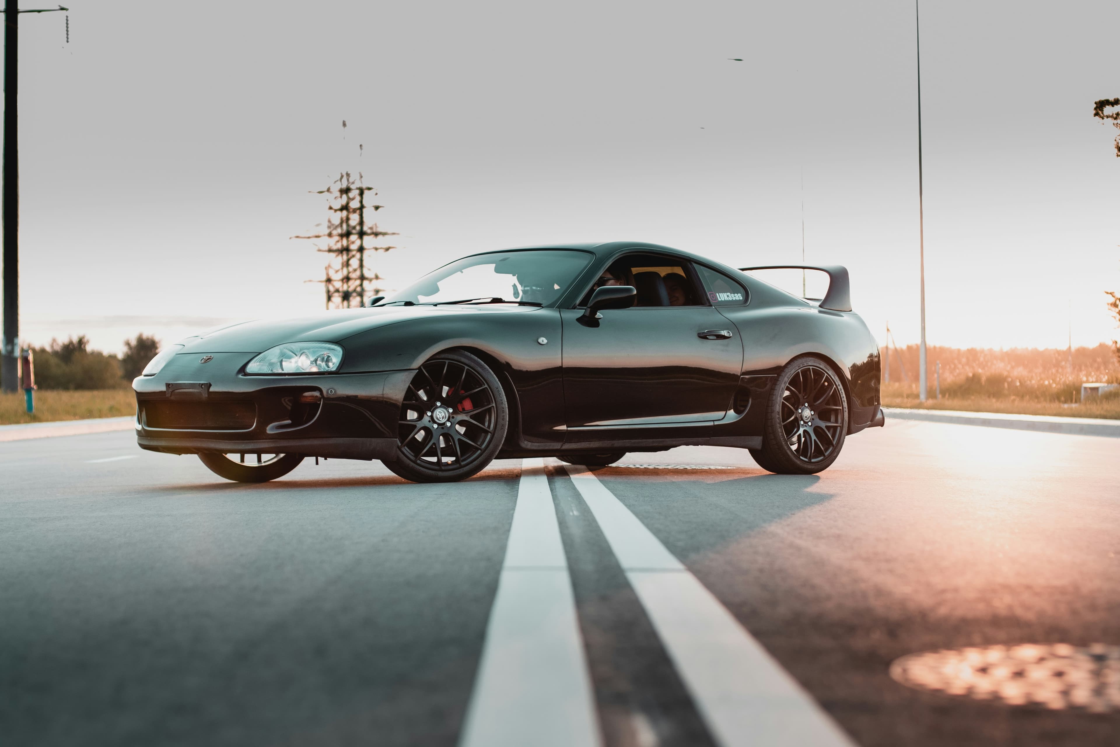 Dark Toyota Supra A80 viewed from the front three-quarter angle in warm evening light, showing its distinctive silhouette and rear wing